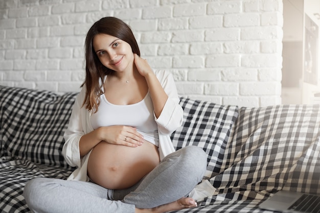 Gratis foto portret van jonge zwangere meisjeszitting op bank die van haar homestay geniet. baby lacht van binnen zoals haar moeder dat doet.