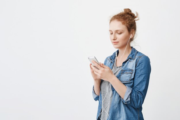 portret van kalme aantrekkelijke roodharige jonge vrouw met haar gekamd in broodje met smartphone en scherm kijken terwijl het luisteren van muziek met een koptelefoon