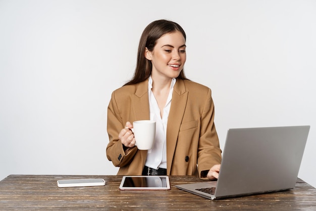 Portret van kantoorvrouw, ceo-ondernemer die op laptop werkt, koffie drinkt en klanten op de website beantwoordt, staande op een witte achtergrond