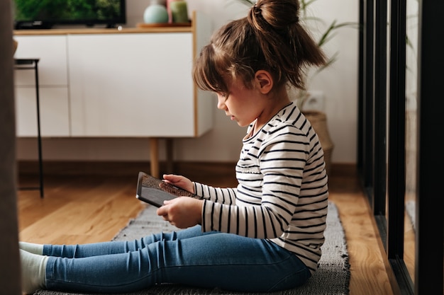 Portret van kleine vrouw in jeans en t-shirt zittend op een tapijt en tablet te houden.