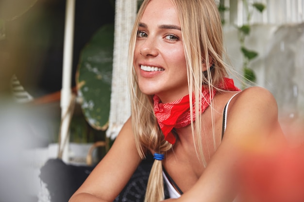 Portret van knap ontspannen jong vrouwelijk model met rode bandana op nek, heeft haar eigen stijl, zit tegen café gezellig interieur