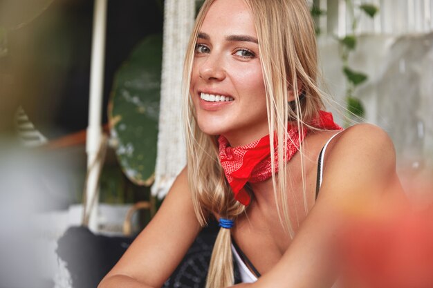 Portret van knap ontspannen jong vrouwelijk model met rode bandana op nek, heeft haar eigen stijl, zit tegen café gezellig interieur