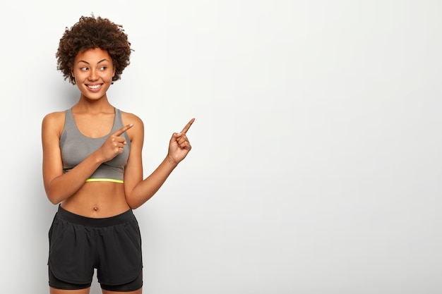 Portret van knappe Afro-vrouw wijst weg op lege ruimte, lacht aangenaam, draagt top en korte broek, kopie ruimte tegen witte muur