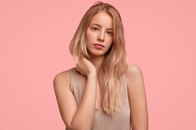 Portret van knappe blonde vrouw raakt nek met handen, ziet er mysterieus en serieus uit, heeft een zuivere huid, gaat buiten lopen, modellen tegen roze muur