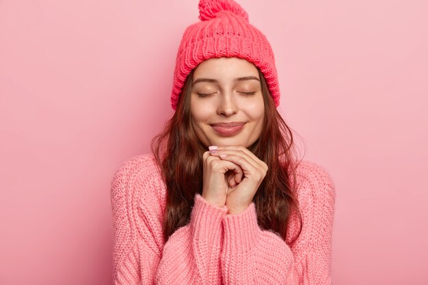 Portret van knappe charmante vrouw houdt beide handen onder de kin, ogen gesloten, diep in gedachten, modellen over roze studiomuur, gekleed in trui en hoed