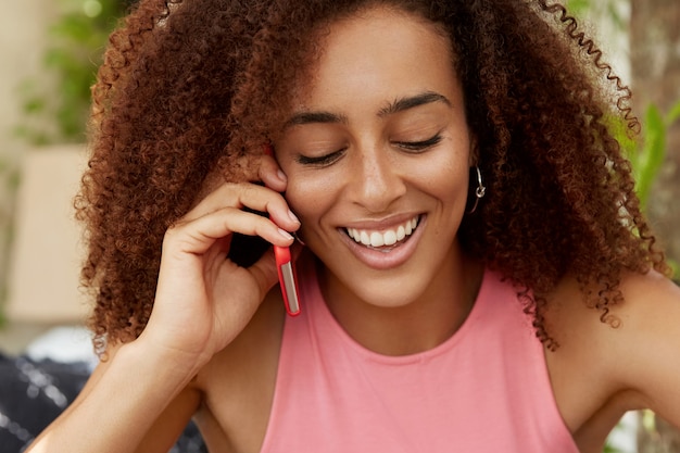 Gratis foto portret van knappe, donkere vrouw met krullend haar, kijkt positief naar beneden, heeft een aangenaam telefoongesprek met beste vriend, deelt indrukken na een goede vakantie in het buitenland