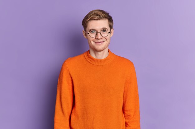 Portret van knappe Europese mannelijke student heeft een zachte glimlach op het gezicht blij om te horen aangenaam nieuws staat opgetogen draagt ronde bril oranje trui