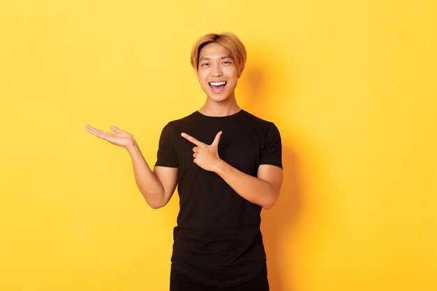 Portret van knappe glimlachende Aziatische kerel die iets bij de hand houdt en vinger ernaar richt, die zich gele muur tevreden bevindt