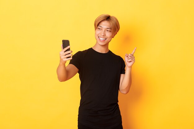 Portret van knappe glimlachende aziatische mens die videogesprek op smartphone heeft en vinger naar iets richt, zich gele muur bevindt