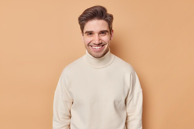 Portret van knappe jonge man met donker haar brede glimlach kijkt graag naar camera draagt casual witte coltrui geïsoleerd over bruine achtergrond Glimlachend volwassen mannelijk model in goed humeur