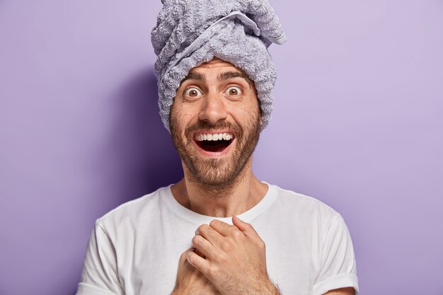 Portret van knappe jonge man met gelukkige uitdrukking, draagt handdoek net na het douchen, verrast om een zachte huid te hebben, gekleed in een casual wit t-shirt