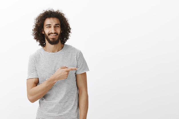 Portret van knappe jongen met charmante glimlach en krullend haar in vrijetijdskleding, naar rechts wijzend en lachend met zelfverzekerde uitdrukking