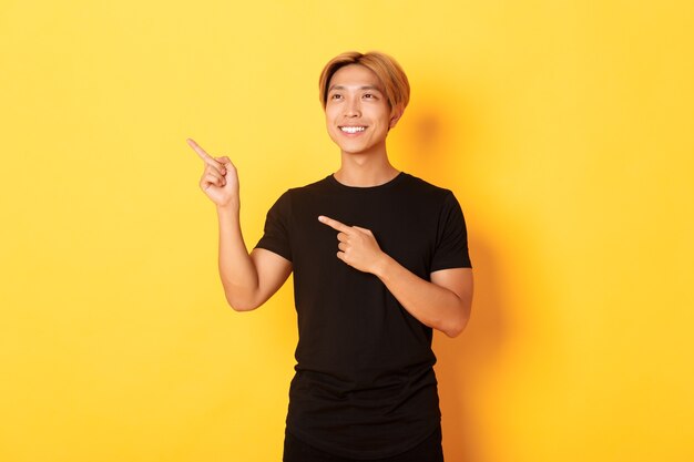 Portret van knappe Koreaanse blonde kerel, wijzende vingers linkerbovenhoek en kijkt tevreden met tevreden glimlach, gele muur.