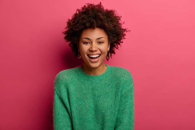 Portret van knappe krullende etnische vrouw glimlacht breed, geniet van vrije dag, heeft een gelukkig gesprek met de gesprekspartner, bespreekt de voorbereiding van de vakantie, draagt een groene trui, geïsoleerd op een roze muur.