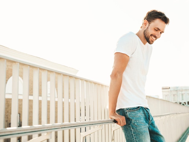 Portret van knappe lachende hipster lamberseksueel modelstijlvolle man gekleed in witte tshirt en spijkerbroek mode man poseren op de straat achtergrond in de buurt van hek op het strand vrolijk en gelukkig