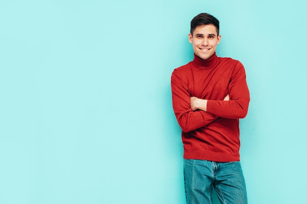 Portret van knappe lachende model Sexy stijlvolle man gekleed in rode coltrui en jeans Fashion hipster man poseren in de buurt van blauwe muur in studio geïsoleerd