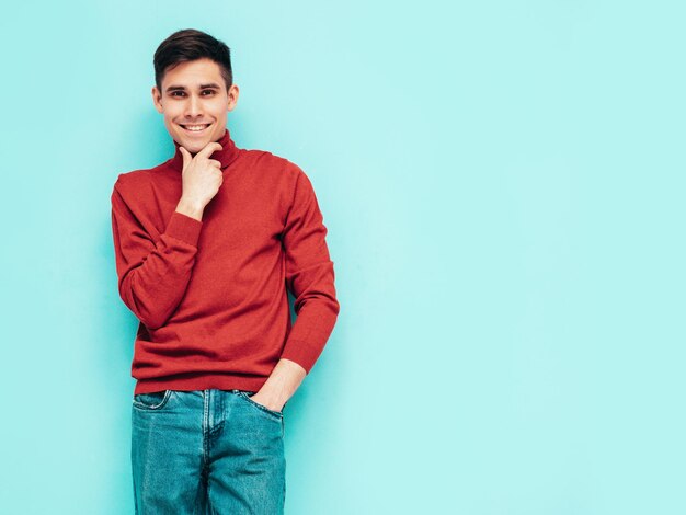 Portret van knappe lachende model Sexy stijlvolle man gekleed in rode coltrui en jeans Fashion hipster man poseren in de buurt van blauwe muur in studio geïsoleerd