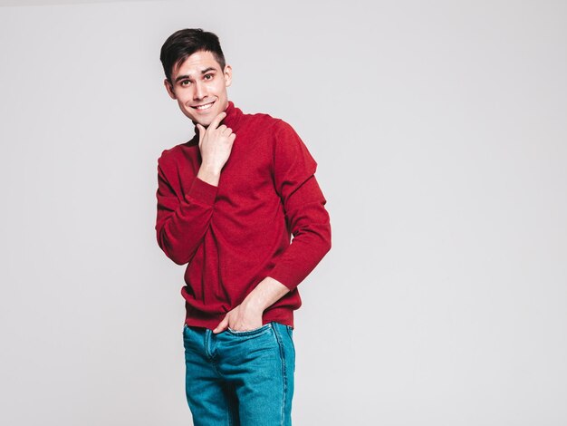 Portret van knappe lachende model Sexy stijlvolle man gekleed in rode coltrui en jeans Fashion hipster man poseren op grijze achtergrond in studio geïsoleerd