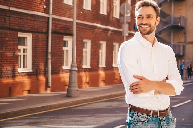 Portret van knappe lachende stijlvolle hipster lamberseksueel model moderne man gekleed in wit overhemd mode man poseren op de straat achtergrond buitenshuis bij zonsondergang gekruiste armen