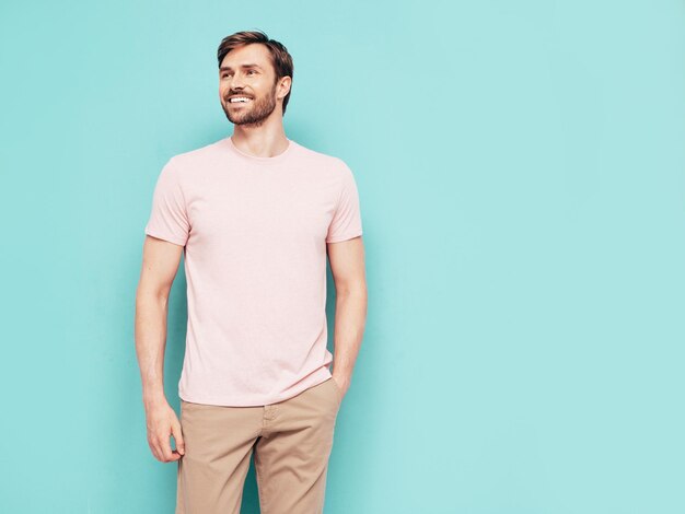 Portret van knappe lachende stijlvolle hipster lamberseksueel model Sexy man gekleed in roze tshirt en broek mode man geïsoleerd op blauwe muur in studio