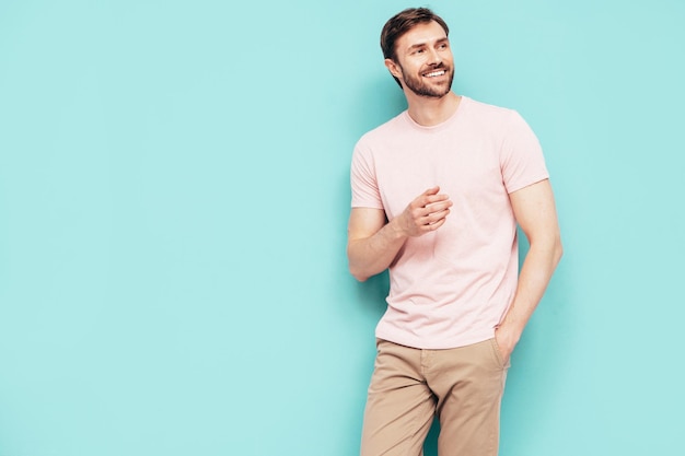 Portret van knappe lachende stijlvolle hipster lamberseksueel model Sexy man gekleed in roze tshirt en broek mode man geïsoleerd op blauwe muur in studio