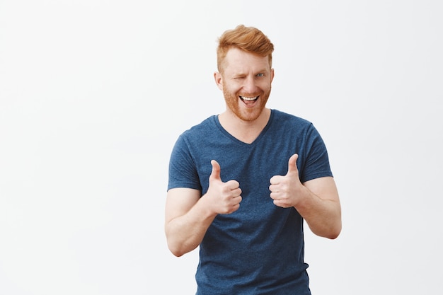 Portret van knappe man met rood haar duimen opdagen en knipogen met hint, leuk vinden en ondersteunen van grote beslissing, juichen over grijze muur