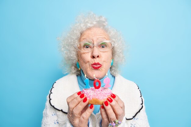 Portret van knappe senior vrouw die kaarsen gaat blazen op donut viert 102e verjaardag ziet er mooi uit heeft rode manicure lichte make-up gekleed in trui