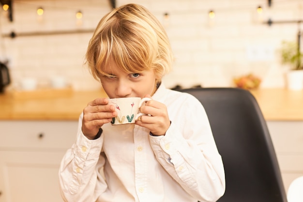 Portret van knappe vrolijke jongen met blond haar, het drinken van thee tijdens het ontbijt voor school, kopje houden en lachend met keukengerei en slinger op aanrecht