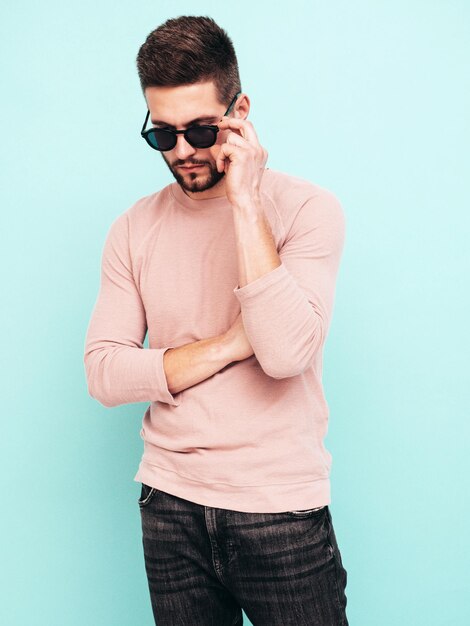 Portret van knappe zelfverzekerde model Sexy stijlvolle man gekleed in roze trui en jeans Fashion hipster man poseren in de buurt van blauwe muur in studio In zonnebril geïsoleerd