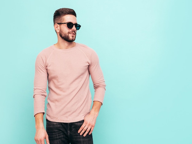 Portret van knappe zelfverzekerde model Sexy stijlvolle man gekleed in roze trui en jeans Fashion hipster man poseren in de buurt van blauwe muur in studio In zonnebril geïsoleerd