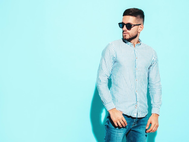 Portret van knappe zelfverzekerde model Sexy stijlvolle man gekleed in shirt en spijkerbroek Fashion hipster man poseren in de buurt van blauwe muur in studio In zonnebril