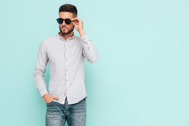 Portret van knappe zelfverzekerde model Sexy stijlvolle man gekleed in shirt en spijkerbroek Mode hipster man poseren in de buurt van blauwe muur in studio vrolijk en gelukkig In zonnebril