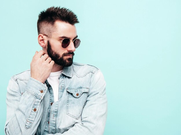 Portret van knappe zelfverzekerde stijlvolle hipster lamberseksueel model man gekleed in jas en spijkerbroek mode man poseren in de buurt van blauwe muur in studio in zonnebril doordachte geïsoleerd