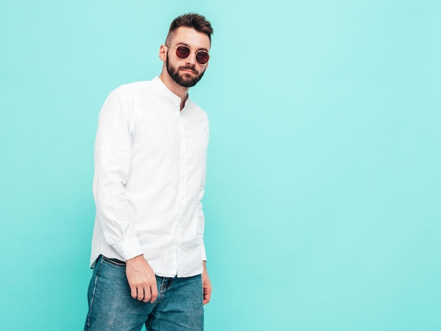 Portret van knappe zelfverzekerde stijlvolle hipster lamberseksueel model Man gekleed in wit overhemd en jeans Mode man geïsoleerd op blauwe muur in studio In zonnebril