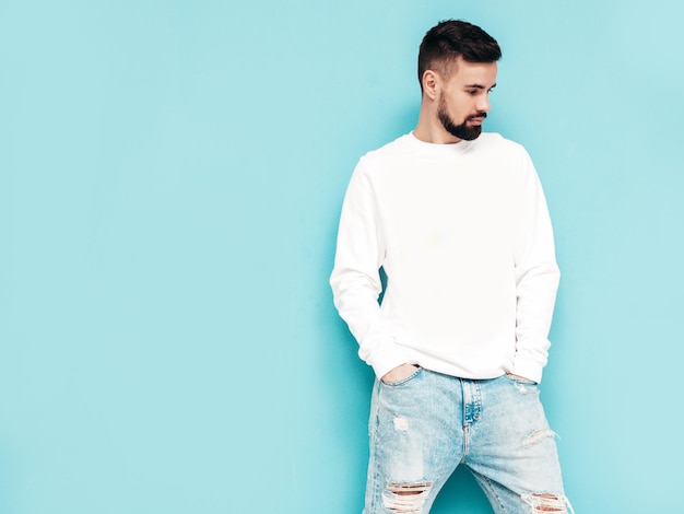 Portret van knappe zelfverzekerde stijlvolle hipster lamberseksueel model Man gekleed in witte trui en jeans Mode man geïsoleerd op blauwe muur in studio