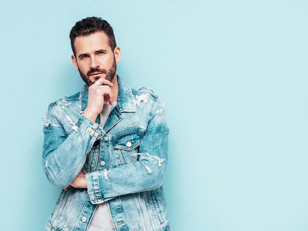 Portret van knappe zelfverzekerde stijlvolle hipster lamberseksueel model Sexy man gekleed in jeans jasje Mode man geïsoleerd op blauwe muur in studio