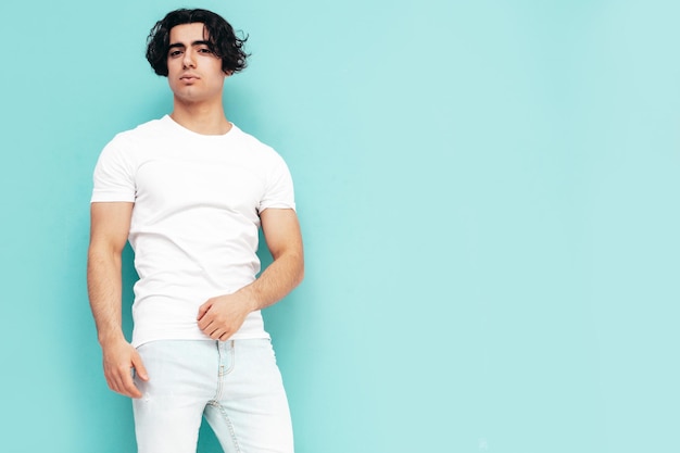 Portret van knappe zelfverzekerde stijlvolle hipster lamberseksueel model Sexy man gekleed in zomer witte T-shirt en jeans kleding Mode man geïsoleerd in studio poseren in de buurt van blauwe muur