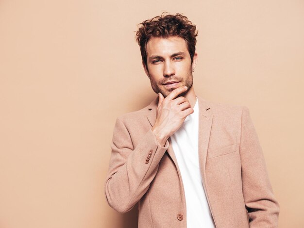 Portret van knappe zelfverzekerde stijlvolle hipster lamberseksueel model Sexy moderne man gekleed in elegant beige pak Mode man met krullend kapsel poseren in studio zijn kin aanraken blauwe ogen