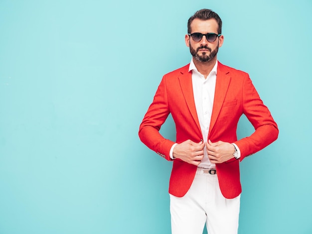 Gratis foto portret van knappe zelfverzekerde stijlvolle hipster lamberseksueel modelsexy moderne man gekleed in elegant rood pak fashion man poseren in studio in de buurt van blauwe muur in zonnebril