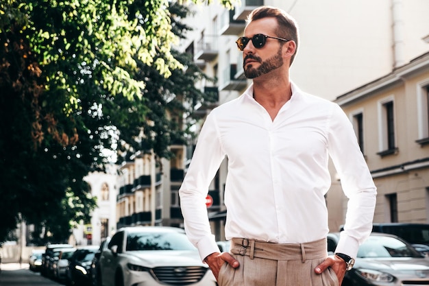 Portret van knappe zelfverzekerde stijlvolle hipster lamberseksueel modelSexy moderne man gekleed in wit overhemd en broek Fashion man poseren op straat achtergrond in Europa stad bij zonsondergang In zonnebril