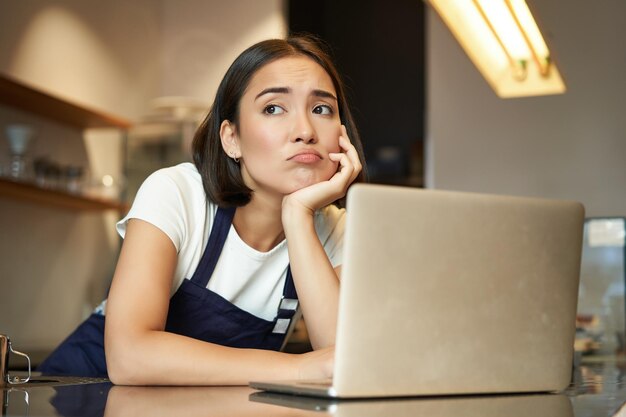 Portret van Koreaanse barista in café die er verdrietig en fronsend grimassend teleurgesteld uitziet terwijl ze aan het werk is in de coffeeshop, leunend op de toonbank in de buurt van laptop