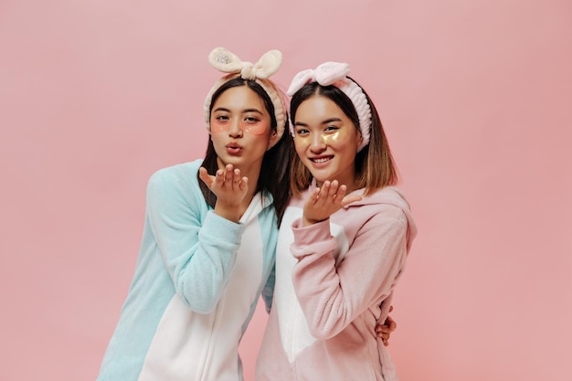 Portret van kortharige Aziatische meisjes in hoofdbanden en met cosmetische ooglapjes Mooie vrouwen in pyjama blazen kus op roze achtergrond