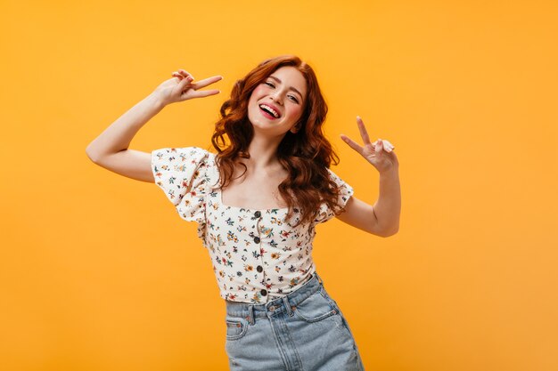 Portret van krullende vrouw in blouse en denimrok die vredesteken op oranje achtergrond tonen.