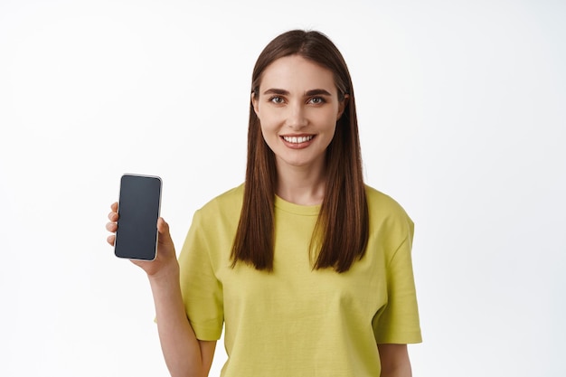 Portret van lachend brunette meisje met smartphonescherm, app-interface of winkel-app, ziet er gelukkig uit, beveelt downloadtoepassing aan, witte achtergrond