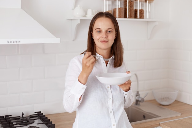 Portret van lachende aantrekkelijke jonge volwassen vrouw met donker haar, gekleed in een wit casual shirt, ontbijten in de keuken, bord in handen houdend, camera kijkend met aangename uitdrukking.