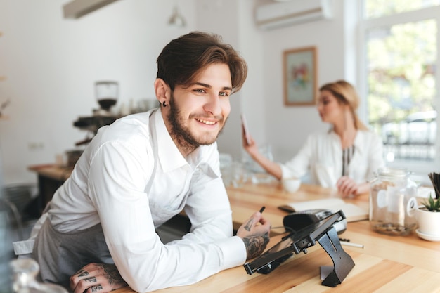 Portret van lachende barista in schort en wit overhemd op werkplek in coffeeshop. Barista kijkt vrolijk opzij terwijl ze aan de balie staat terwijl mooi meisje op de achtergrond haar mobiele telefoon gebruikt in café