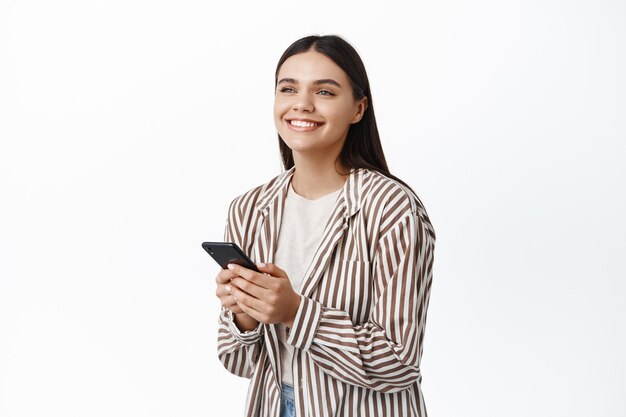 Portret van lachende blanke vrouw in trendy outfit, chatten of app gebruiken, opzij kijken gelukkig, staande tegen een witte muur