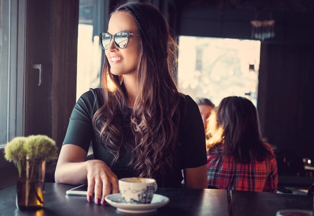 Portret van lachende brunette vrouw in zonnebril, drinkt koffie in de ochtend in een café.