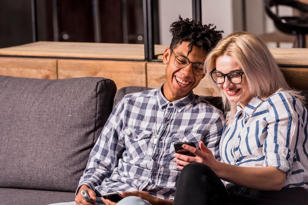 Portret van lachende jonge paar zittend op de bank kijken naar mobiele telefoon