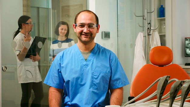 Portret van lachende man verpleegster in tandartspraktijk terwijl pediatrische arts praat met de patiënt op de achtergrond. Stomatoloog assistent kijken camera zittend op stoel in stomatologische kliniek.